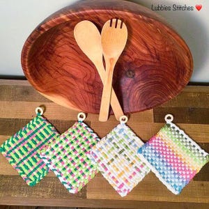 May include: A wooden salad bowl with two wooden serving spoons and four woven pot holders. The pot holders are square-shaped and feature a variety of colors, including green, pink, white, and blue. The text "Lubbies Stitches" is visible.