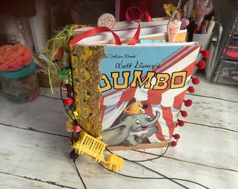 May include: A handmade Dumbo-themed journal. The cover features the classic Dumbo illustration with the title in bold yellow letters. The journal is embellished with gold trim, red pom-poms, and decorative beads. Red ribbon handles are attached to the top.