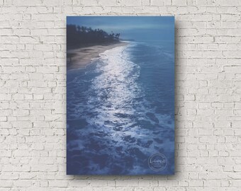 May include: A blue and white photograph of a beach with waves crashing on the shore. The photo was taken from a high vantage point, looking down at the beach. The water is a deep blue, and the waves are white and foamy. The beach is sandy and there are trees in the background.