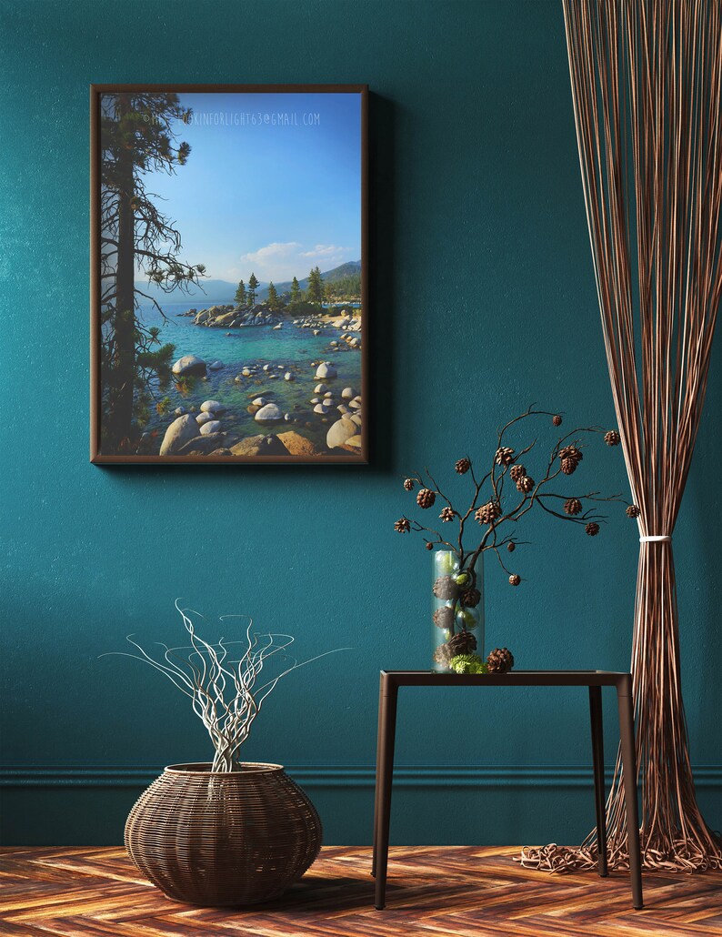 May include: A framed photograph of a lake with a tree in the foreground and mountains in the background. The lake is a light blue color and the mountains are a dark blue color. The tree is a dark green color and the sky is a light blue color. The photo is hanging on a teal wall with a brown wicker basket and a small table with a vase of dried branches.