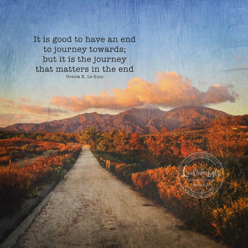 May include: A quote by Ursula K. Le Guin about the importance of the journey, set against a scenic backdrop of a winding path through a field of autumn foliage, leading towards a mountain range under a cloudy sky.