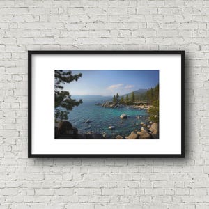 May include: A framed photograph of a scenic lake with a rocky shoreline and a pine tree in the foreground. The water is a vibrant blue and the sky is a clear blue with a few white clouds. The mountains in the distance are covered in trees.