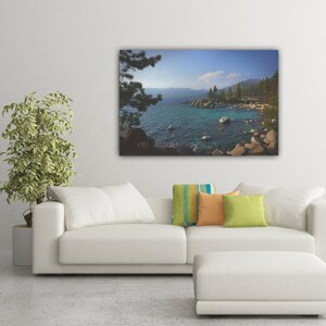 May include: A scenic photograph of a lake with a rocky shoreline and lush green trees. The water is a vibrant blue and the sky is a clear blue with a few white clouds.