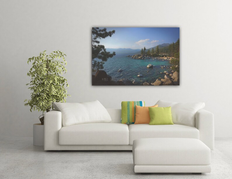 May include: A scenic photograph of a lake with a rocky shoreline and lush green trees. The water is a vibrant blue and the sky is a clear blue with a few white clouds.
