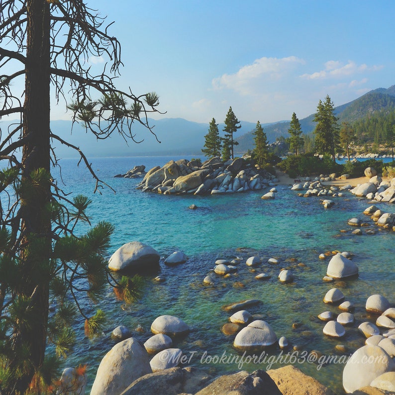 May include: A scenic view of a lake with turquoise water and large, smooth rocks scattered throughout. The water is calm and clear, reflecting the blue sky and white clouds.  A few pine trees are visible on the shore, adding to the natural beauty of the scene.