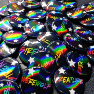 Può includere: Una collezione di bottoni rotondi iridescenti con varie parole e frasi in testo colorato, tra cui "SIRE", "WEREWOLF" e "MONSTER". Ogni bottone presenta un design a stella bianca.