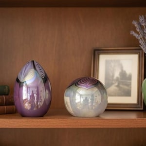 May include: Two decorative glass vases on a wooden shelf. One is a teardrop shape in shades of purple and black, the other is a round, iridescent glass. A framed picture and a green vase with lavender are also on the shelf.