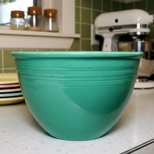 May include: A turquoise ceramic mixing bowl with horizontal ridges. The bowl is set on a white countertop, with a white stand mixer and jars in the background. The bowl is a classic kitchenware item.