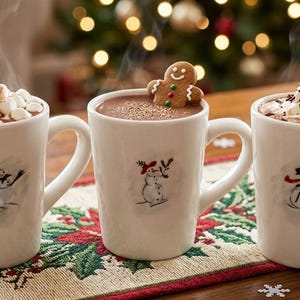 May include: Three white ceramic mugs filled with hot cocoa, each featuring a unique snowman illustration. The first mug is topped with marshmallows, the second with a gingerbread cookie, and the third with whipped cream and a candy cane.