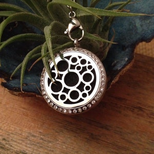 May include: A silver aromatherapy pendant with a circular design featuring multiple open circles. The pendant is adorned with small, clear crystals around the edge and hangs from a silver chain with a clasp. The pendant is displayed with green plant life.