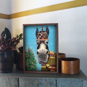May include: A framed illustration of a donkey wearing a colorful knitted hat and scarf, with a small Christmas tree in the background. The donkey has a blue background with white snowflakes.