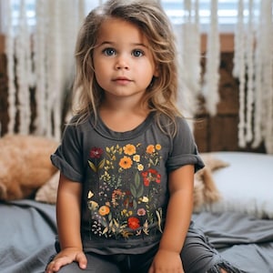 May include: A young girl wearing a gray t-shirt with a floral print. The shirt features a variety of flowers in shades of orange, yellow, red, and pink. The girl is sitting on a gray bed with her legs crossed.