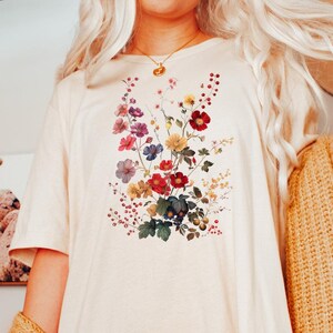 May include: A white oversized t-shirt with a floral print featuring a variety of colorful flowers. The shirt is worn by a person with long blonde hair and a brown cardigan.