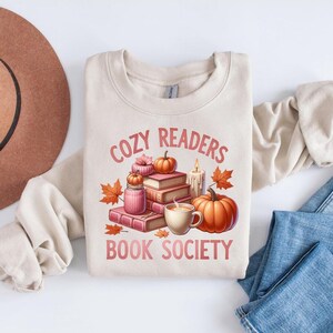 May include: A beige sweatshirt with the text "Cozy Readers Book Society" in pink lettering. The design features a stack of books, a mug of coffee, pumpkins, and fall leaves.