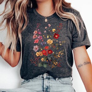 May include: A dark gray t-shirt with a floral print featuring red, yellow, pink, and purple flowers. The flowers are arranged in a bouquet shape and are surrounded by green leaves. The t-shirt is being worn by a person sitting on a stool.