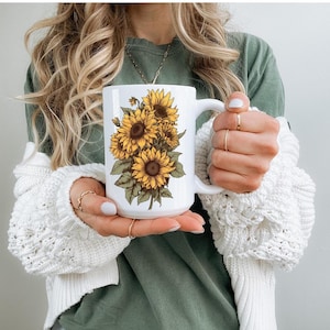Pode incluir: Caneca de cerâmica branca com um gráfico de um buquê de girassóis amarelos. A caneca tem uma alça preta.