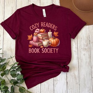 May include: Burgundy t-shirt with a graphic of a stack of books, a lit candle, a mug of hot beverage, and pumpkins. The text "Cozy Readers Book Society" is printed on the shirt.