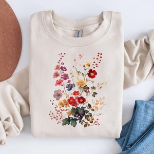 May include: A beige sweatshirt with a floral print featuring a variety of colorful flowers, including red, yellow, orange, purple, and pink.