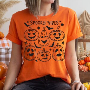 Puede incluir: Camiseta naranja con un gráfico negro de calabazas sonrientes y el texto "Spooky Vibes".