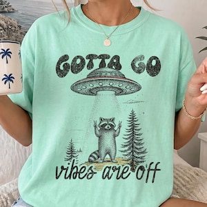 May include: A mint green t-shirt featuring a graphic print of a raccoon standing between two pine trees, making a peace sign gesture. Above the raccoon is a flying saucer with a beam of light, and the text reads GOTTA GO vibes are off.