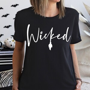 Puede incluir: Camiseta negra con la palabra "Wicked" en escritura blanca, con un diseño de escoba de bruja. Una prenda con temática de Halloween, perfecta para la temporada de miedo.