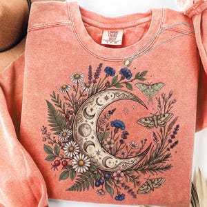May include: A coral-colored crewneck sweatshirt featuring a graphic print of a crescent moon surrounded by various wildflowers, ferns, and three moths. The inner neck label displays the text Comfort Colors. The fabric has a washed, textured appearance.