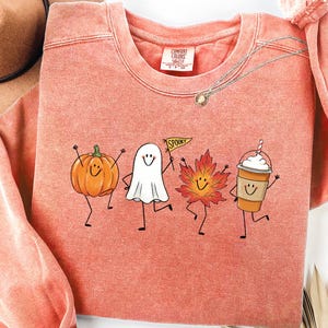 May include: A coral-colored crewneck sweatshirt featuring a graphic print of four animated characters: an orange pumpkin, a white ghost holding a pennant that reads SPOOKY, a red autumn leaf, and a coffee cup with whipped cream, all with smiling faces.
