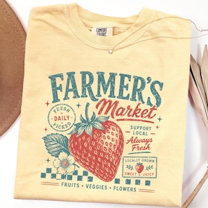 May include: A pale yellow crew neck t-shirt featuring a graphic print with the text FARMER'S MARKET, a large red strawberry, and smaller text including FRESH DAILY PICKED, SUPPORT LOCAL, ALWAYS FRESH, LOCALLY GROWN, SWEET & JUICY, FRUITS, VEGGIES, FLOWERS.