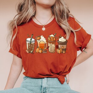 Könnte beinhalten: Eine Frau trägt ein rostfarbenes T-Shirt mit einem Aufdruck von vier Getränken mit Kürbisgewürz. Das Shirt ist in der Taille gebunden und die Frau trägt eine blaue Jeans.