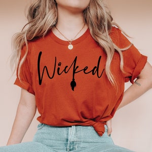 Puede incluir: Una mujer lleva una camiseta de color óxido con el texto "Wicked" en escritura negra con una escoba de bruja y un fantasma gráfico. Lleva pantalones vaqueros azules y zapatillas blancas.