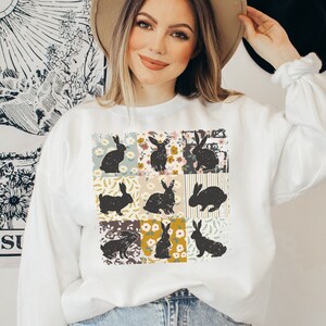 May include: White sweatshirt with a patchwork design featuring black bunny silhouettes and floral patterns.