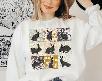 May include: White sweatshirt with a patchwork design featuring black bunny silhouettes and floral patterns.