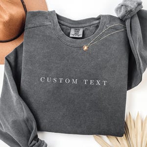 May include: A charcoal gray crewneck sweatshirt featuring the words CUSTOM TEXT embroidered in white thread across the chest. The garment has a vintage-washed appearance, a ribbed collar, and a Comfort Colors brand label inside the neckline.