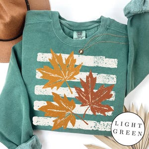 May include: A sage green crewneck sweatshirt featuring a graphic of three maple leaves in shades of orange and brown layered over horizontal white distressed stripes. The interior neck label reads Comfort Colors with size S.