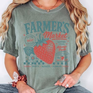 May include: A sage green short-sleeve t-shirt featuring a graphic print of a red strawberry and the text Farmer's Market, Fresh Daily Picked, Support Local, Always Fresh, Locally Grown, Sweet & Juicy, and Fruits, Veggies, Flowers in red and teal.