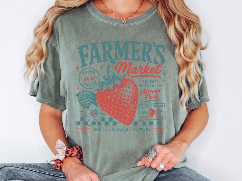 May include: A sage green short-sleeve t-shirt featuring a graphic print of a red strawberry and the text Farmer's Market, Fresh Daily Picked, Support Local, Always Fresh, Locally Grown, Sweet & Juicy, and Fruits, Veggies, Flowers in red and teal.