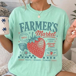 May include: A mint green t-shirt featuring a graphic print that reads "FARMER'S Market" in blue and red text. The design includes a large red strawberry, a small flower, and the phrases "FRESH DAILY PICKED," "SUPPORT LOCAL," "ALWAYS FRESH," "LOCALLY GROWN," "SWEET & JUICY," and "FRUITS VEGGIES FLOWERS."