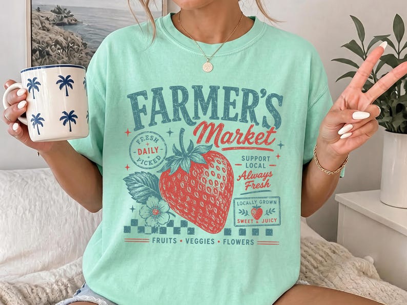 May include: A mint green t-shirt featuring a graphic print that reads "FARMER'S Market" in blue and red text. The design includes a large red strawberry, a small flower, and the phrases "FRESH DAILY PICKED," "SUPPORT LOCAL," "ALWAYS FRESH," "LOCALLY GROWN," "SWEET & JUICY," and "FRUITS VEGGIES FLOWERS."