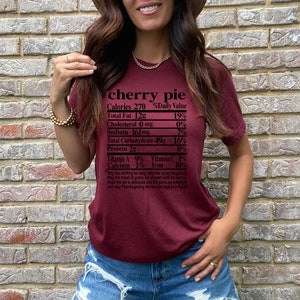 May include: Burgundy-colored t-shirt with the words "cherry pie" and a nutrition facts label printed on the front. The label lists calories, fat, cholesterol, sodium, carbohydrates, and protein content. A Thanksgiving-themed poem is printed below.