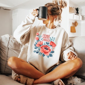 Könnte beinhalten: Ein weißer Pullover mit einem Blumendesign und dem Text "CAPABLE & WORTHY ENOUGH" in roten Buchstaben.