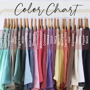 May include: A color chart displaying a row of t-shirts hanging on a gold metal rack. The shirts are labeled with text including Chalky Mint, Island Reef, Seafoam, Bay, Khaki, Light Green, Moss, Chambray, Blue Jean, Blossom, Crunchberry, Watermelon, Crimson, Orchid, Violet, Berry, Banana, Mustard, Terracotta, Yam, Espresso, Ivory, White, Pepper, and Black.