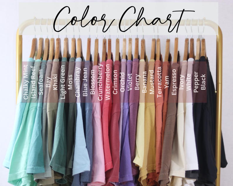May include: A color chart displaying a row of t-shirts hanging on a gold metal rack. The shirts are labeled with text including Chalky Mint, Island Reef, Seafoam, Bay, Khaki, Light Green, Moss, Chambray, Blue Jean, Blossom, Crunchberry, Watermelon, Crimson, Orchid, Violet, Berry, Banana, Mustard, Terracotta, Yam, Espresso, Ivory, White, Pepper, and Black.