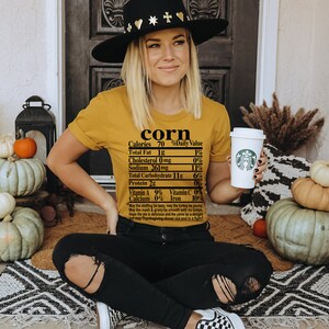 May include: Mustard yellow t-shirt with the word "corn" and a nutrition label printed on it. The person is wearing a black hat and black jeans. Pumpkins and fall decor are in the background.