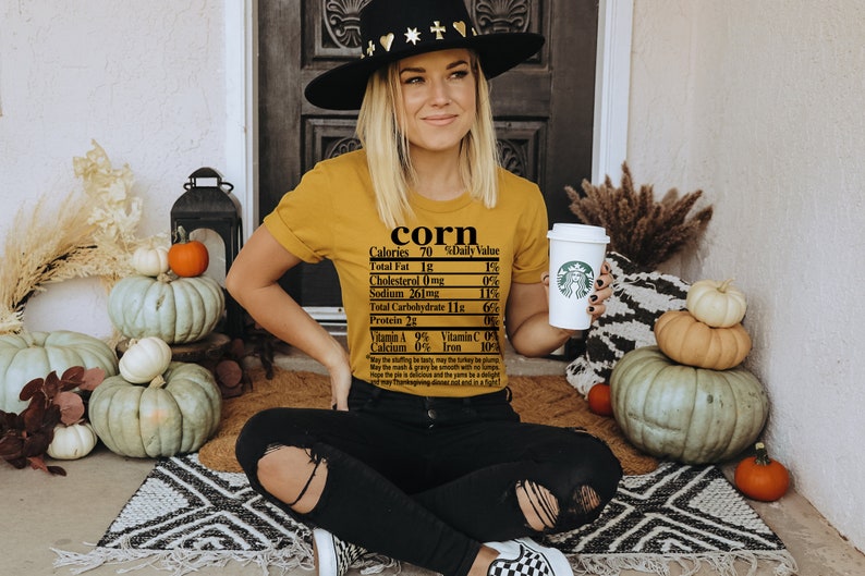 May include: Mustard yellow t-shirt with the word "corn" and a nutrition label printed on it. The person is wearing a black hat and black jeans. Pumpkins and fall decor are in the background.