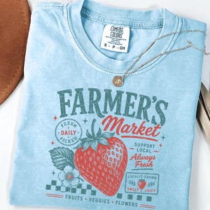 May include: A light blue short-sleeve t-shirt featuring a graphic print that reads "FARMER'S MARKET" with a large red strawberry, a small flower, and text including "FRESH DAILY PICKED," "SUPPORT LOCAL," "Always Fresh," and "LOCALLY GROWN SWEET & JUICY."