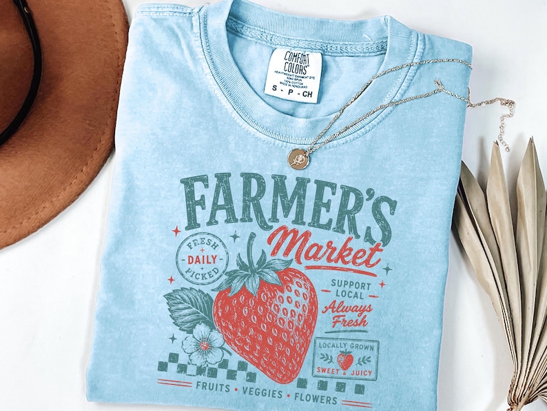 May include: A light blue short-sleeve t-shirt featuring a graphic print that reads "FARMER'S MARKET" with a large red strawberry, a small flower, and text including "FRESH DAILY PICKED," "SUPPORT LOCAL," "Always Fresh," and "LOCALLY GROWN SWEET & JUICY."