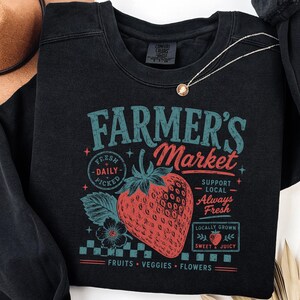 May include: A black crewneck sweatshirt featuring a graphic print with the text Farmer's Market, Fresh Daily Picked, Support Local Always Fresh, Locally Grown Sweet & Juicy, and Fruits Veggies Flowers, centered around a large red strawberry illustration.