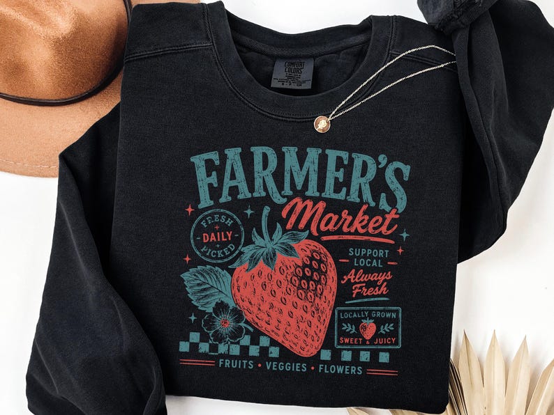 May include: A black crewneck sweatshirt featuring a graphic print with the text Farmer's Market, Fresh Daily Picked, Support Local Always Fresh, Locally Grown Sweet & Juicy, and Fruits Veggies Flowers, centered around a large red strawberry illustration.