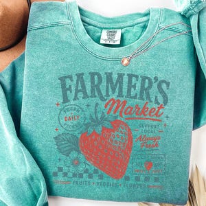 May include: A teal crewneck sweatshirt featuring a graphic print of a large red strawberry and the text Farmer's Market, Fresh Daily Picks, Support Local, Always Fresh, and Fruits Veggies Flowers. The garment has a Comfort Colors brand label.