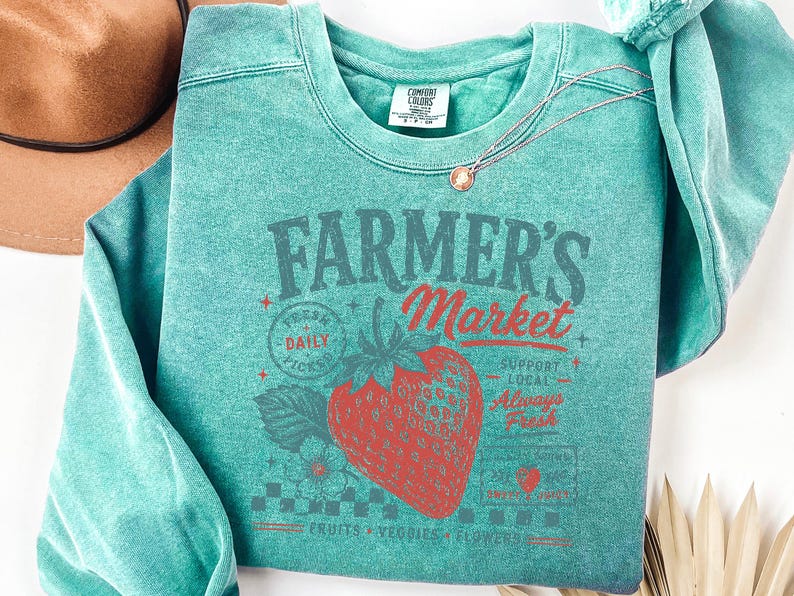 May include: A teal crewneck sweatshirt featuring a graphic print of a large red strawberry and the text Farmer's Market, Fresh Daily Picks, Support Local, Always Fresh, and Fruits Veggies Flowers. The garment has a Comfort Colors brand label.